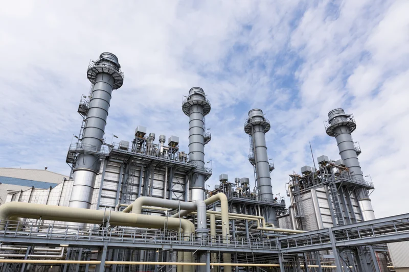 Industrial gas turbine power plant with multiple tall metal smokestacks and interconnected pipes under a partly cloudy sky