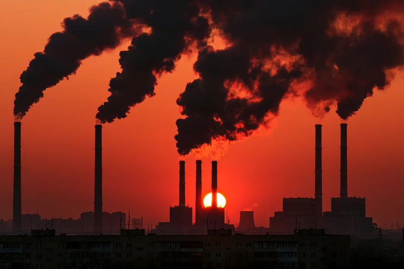 Industrial power plant smokestacks emitting dark smoke against orange sunset sky