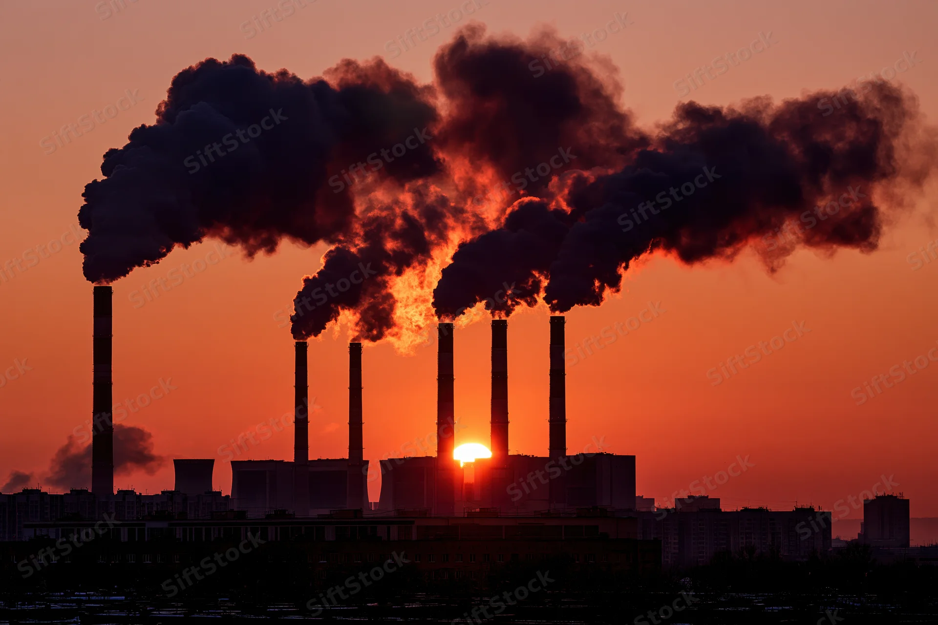 Industrial power plant with multiple smokestacks emitting dark smoke at sunset over city silhouette