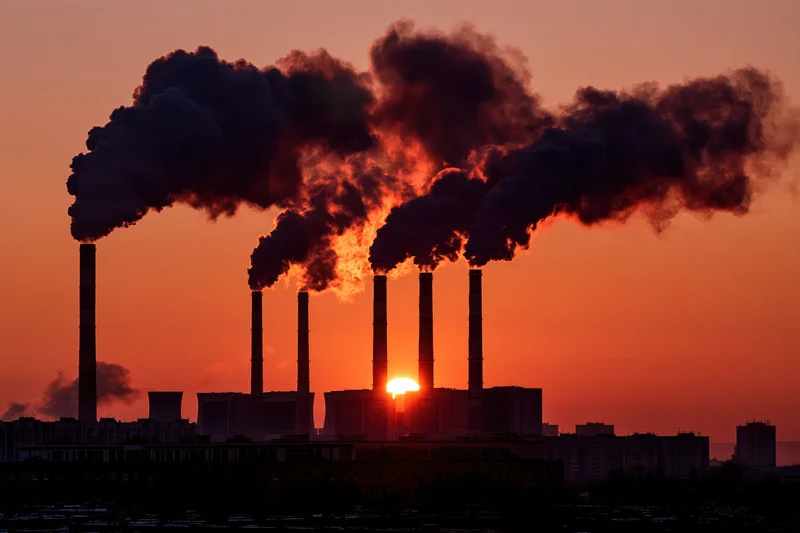 Industrial power plant with multiple smokestacks emitting dark smoke at sunset over city silhouette