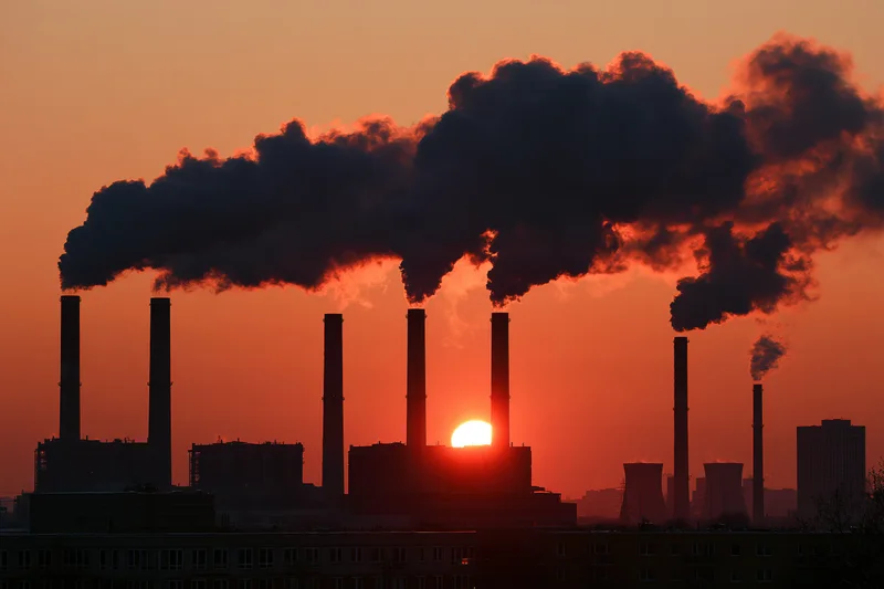 Industrial power plant with multiple smokestacks emitting dark smoke at sunset over city buildings