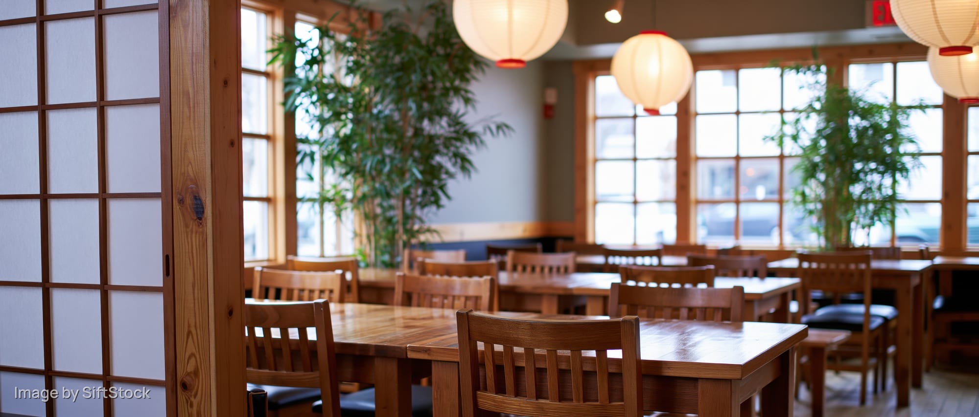 Empty japanese restaurant interior with wooden tables and chairs, shoji screens, hanging paper lanterns, and large windows letting in natural light