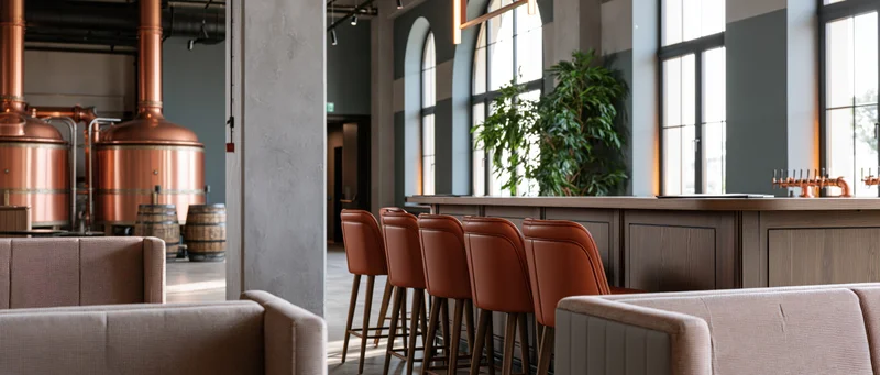 Empty craft brewery taproom interior with copper brewing tanks, wooden bar, leather stools and large windows