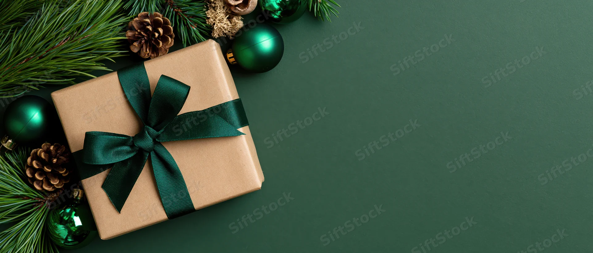 Kraft paper gift box tied with dark green ribbon arranged with pine branches, pinecones and green ornaments on dark green surface