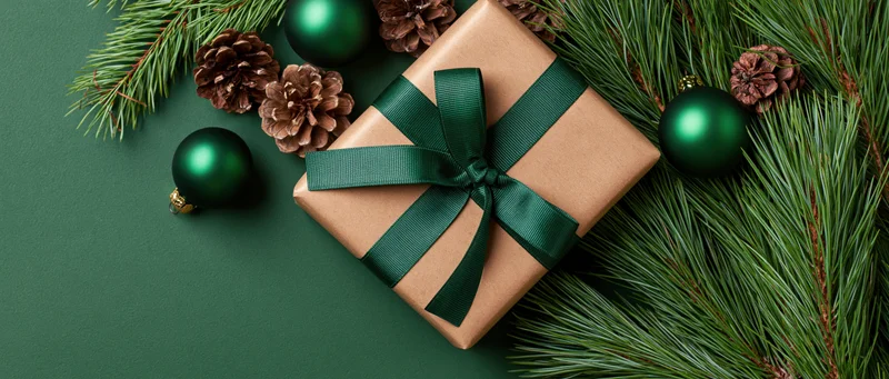 Gift box wrapped in kraft paper with dark green ribbon surrounded by pine branches and ornaments on green background