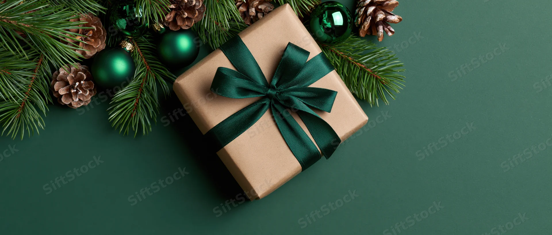 Kraft paper gift box tied with dark green ribbon surrounded by pine branches and ornaments on green background