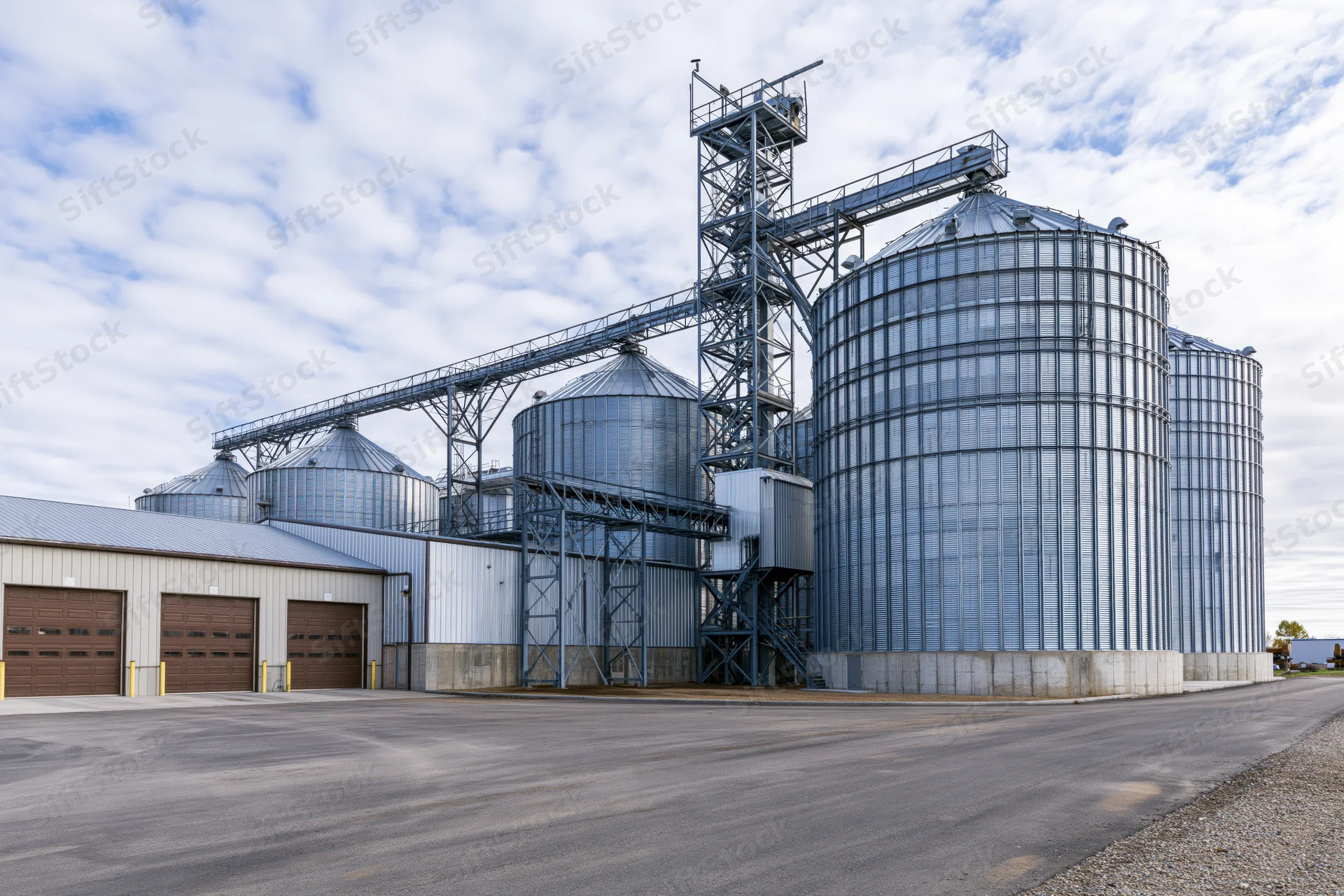 Large agricultural grain elevator complex with multiple tall metal silos and conveyor system under partly cloudy sky