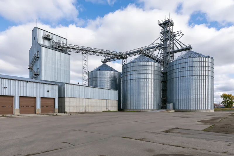 Large agricultural grain elevator complex with multiple tall metal silos and conveyor framework under a partly cloudy sky