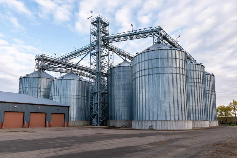 Large agricultural grain elevator complex with multiple tall metal silos and conveyor framework under a partly cloudy sky