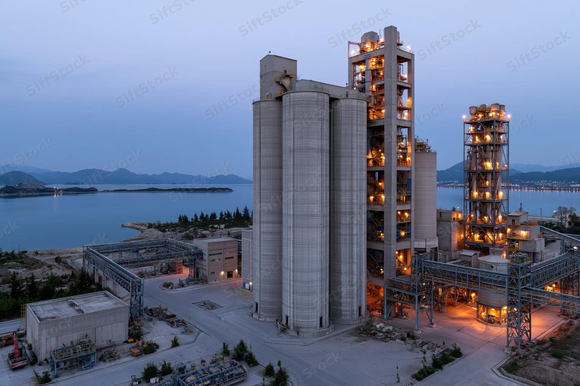 Large cement factory complex with tall concrete silos and illuminated towers at twilight near coastal water