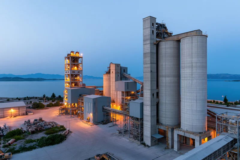 Cement factory complex with tall concrete silos and industrial structures at twilight near water