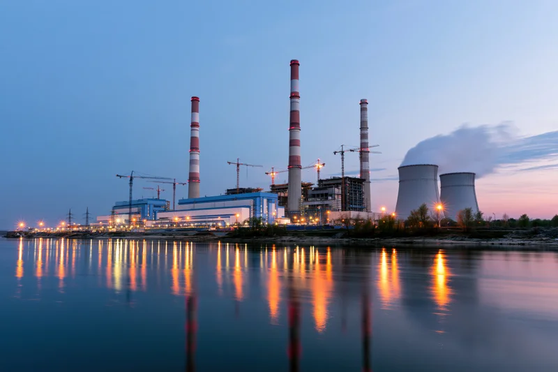 Coal-fired power plant under construction at dusk with cooling towers and smokestacks reflected in river water
