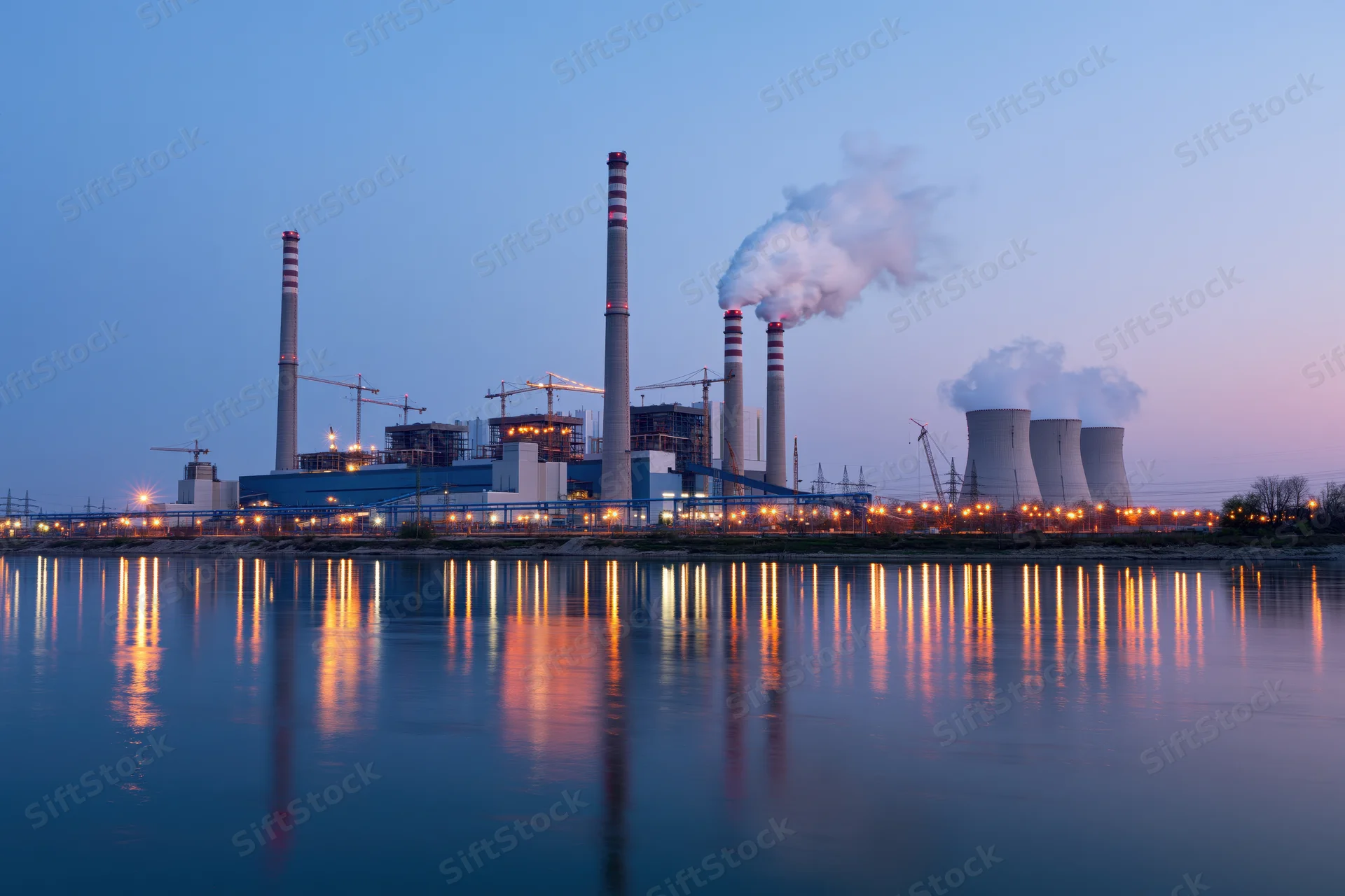 Coal-fired power plant under construction at dusk with smoke rising from chimneys and lights reflecting on the river