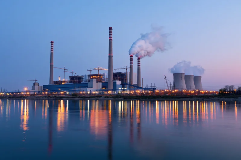 Coal-fired power plant under construction at dusk with smoke rising from chimneys and lights reflecting on the river