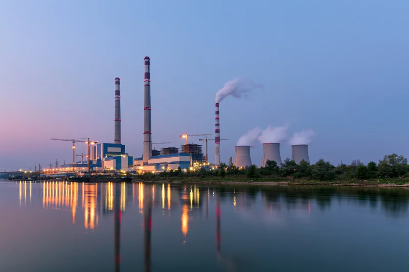 Coal-fired power plant under construction at dusk with smokestacks and cooling towers reflected in river water