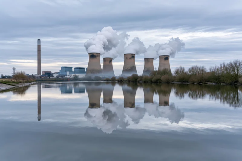 Coal-fired power plant with four cooling towers emitting steam, reflected in calm water
