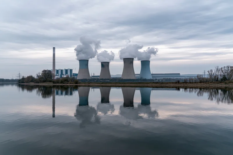 Coal-fired power plant with four cooling towers emitting steam, reflected in calm river water under overcast sky