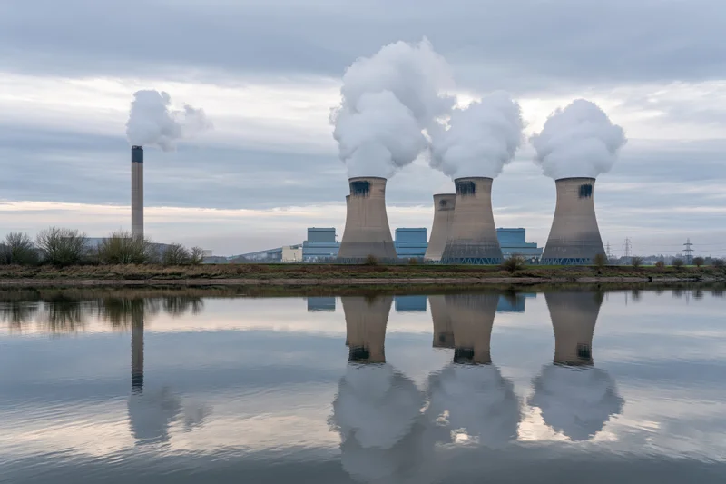 Coal-fired power plant with four hyperbolic cooling towers emitting steam, reflected in calm river water under overcast sky