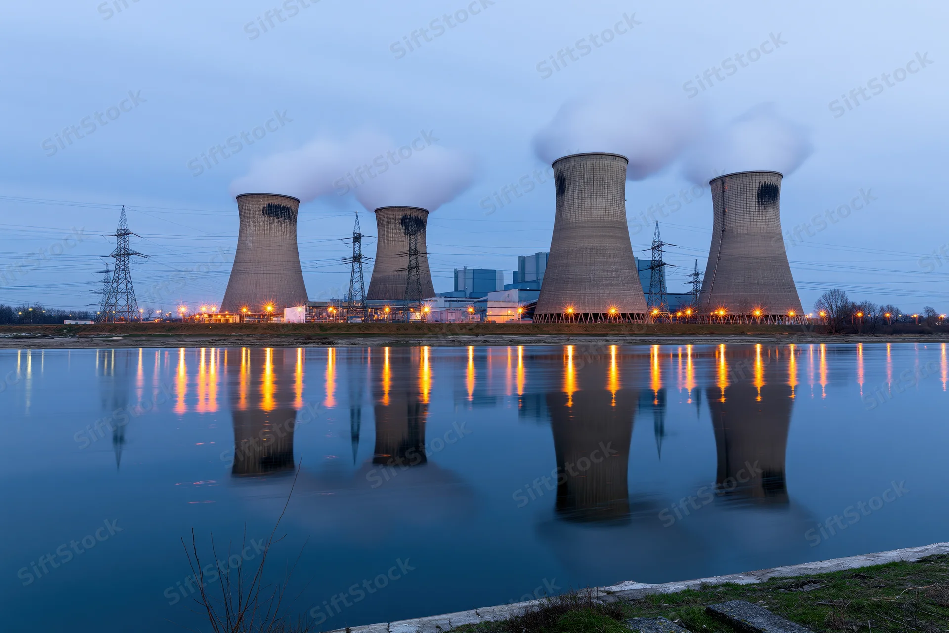 Four cooling towers emitting steam at a coal power plant reflected in calm water during twilight