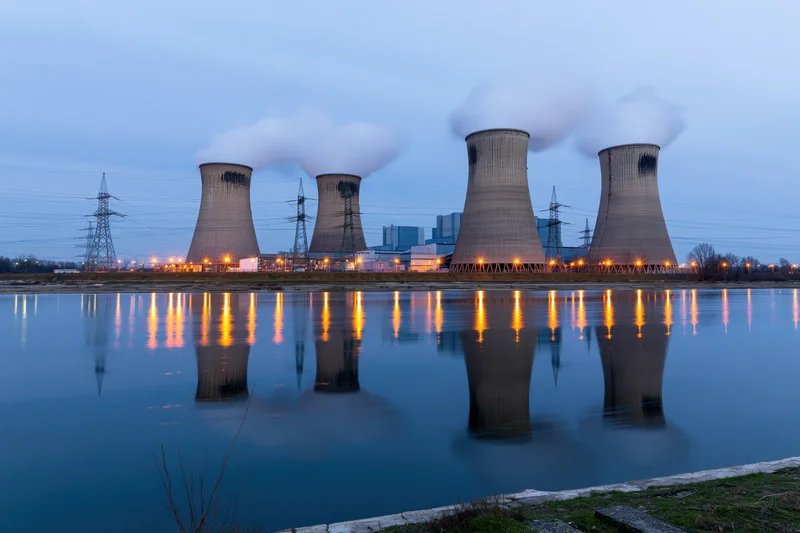 Four cooling towers emitting steam at a coal power plant reflected in calm water during twilight