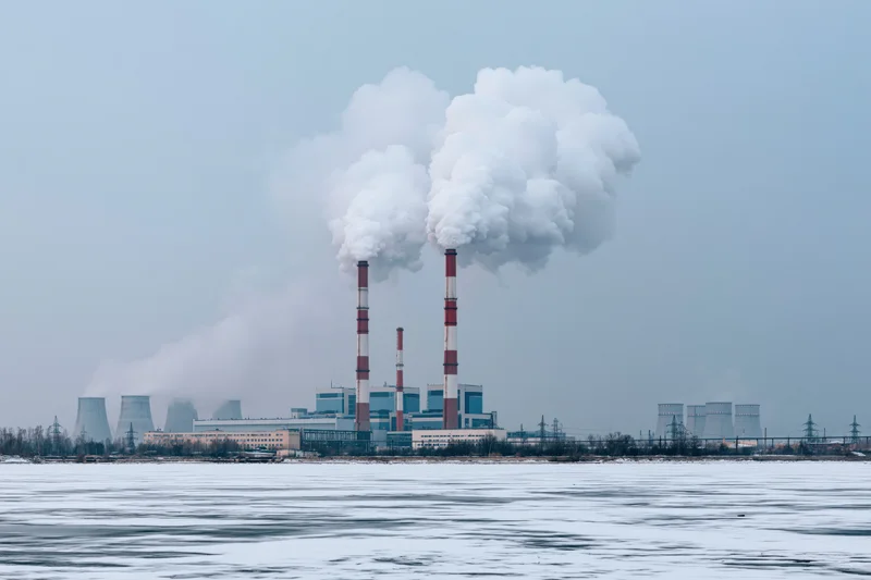 Coal power plant emitting smoke from tall chimneys across frozen river