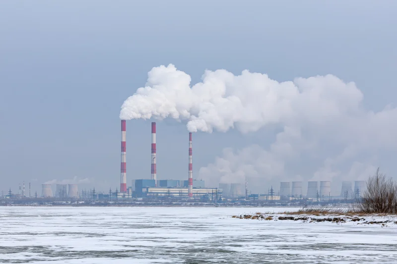 Coal power plant emitting smoke from tall red-and-white striped chimneys across a frozen river in winter