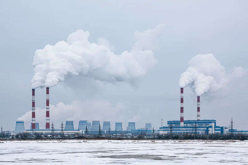Coal power plant with red-and-white striped smokestacks emitting steam in snowy winter landscape