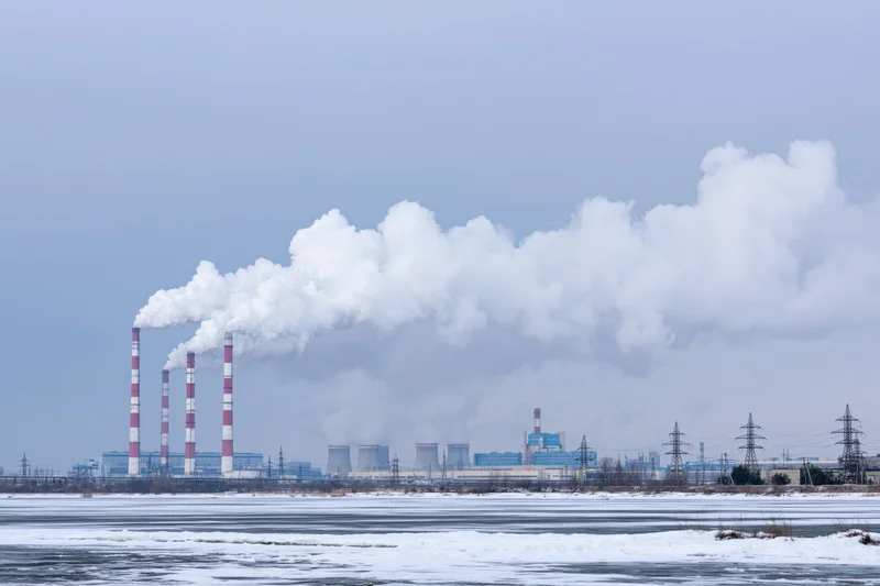 Coal power plant emitting smoke from tall red-and-white striped chimneys beside a frozen river