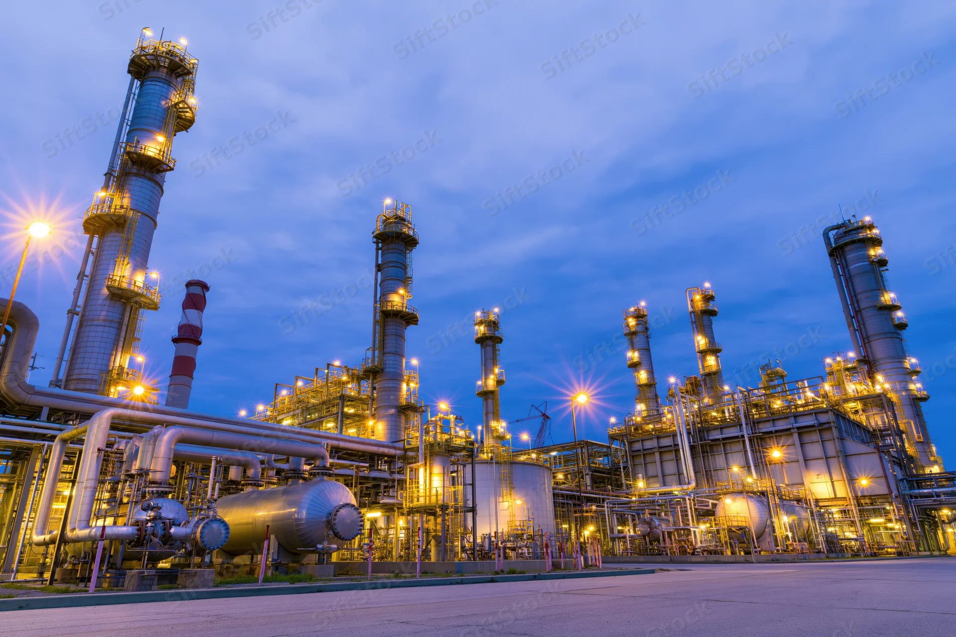 Oil refinery complex at twilight with illuminated towers and pipelines under blue sky