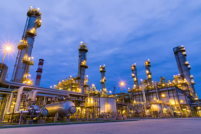Oil refinery complex at twilight with illuminated towers and pipelines under blue sky