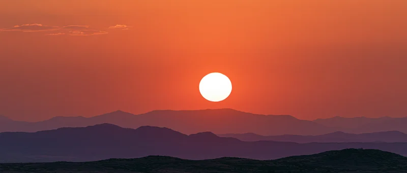 Large glowing sun at dusk above layered mountain silhouettes in a desert landscape