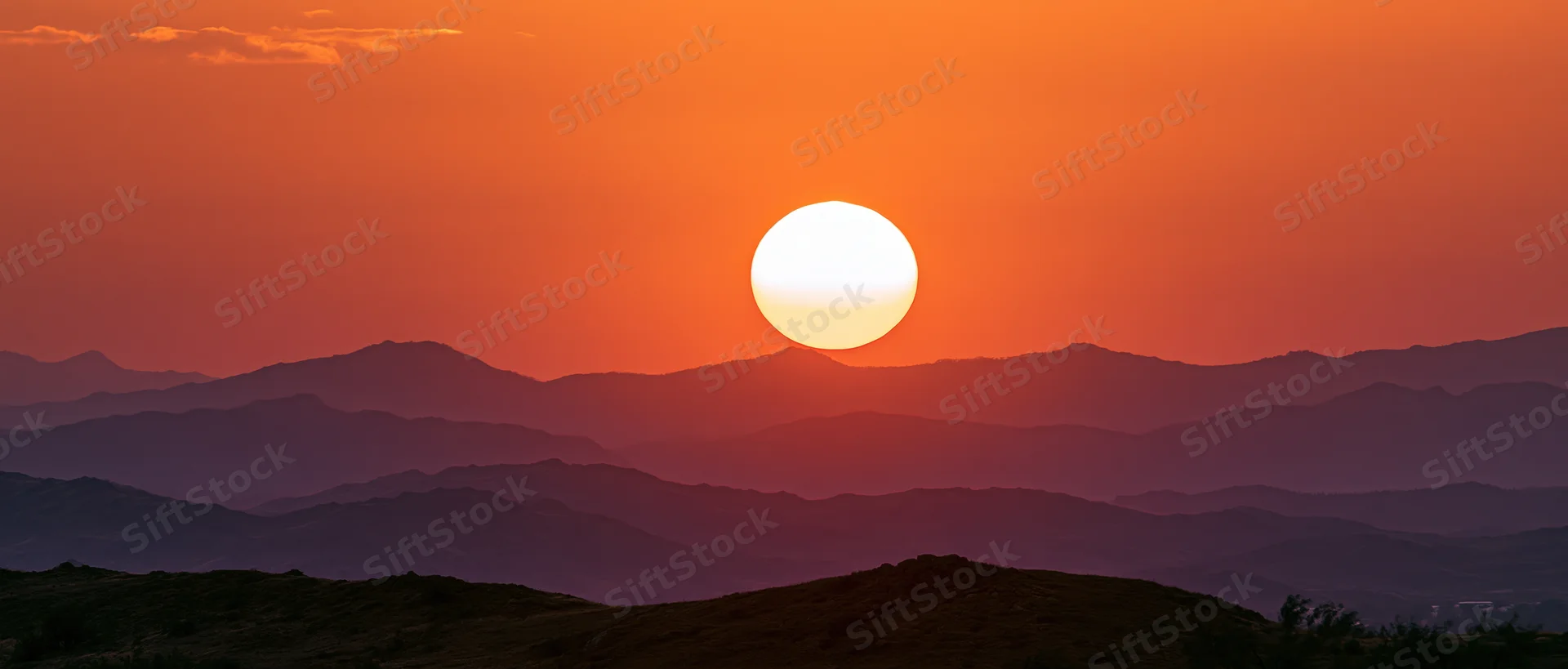 Large glowing sun at dusk above layered mountain silhouettes in a vast landscape