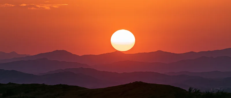 Large glowing sun at dusk above layered mountain silhouettes in a vast landscape