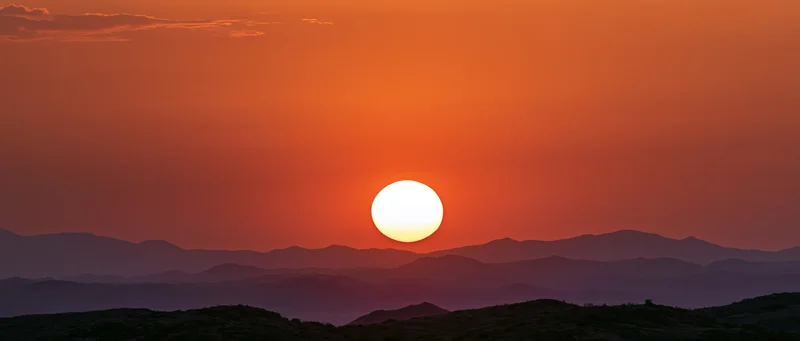 Large glowing sun at dusk above layered mountain silhouettes in a desert landscape