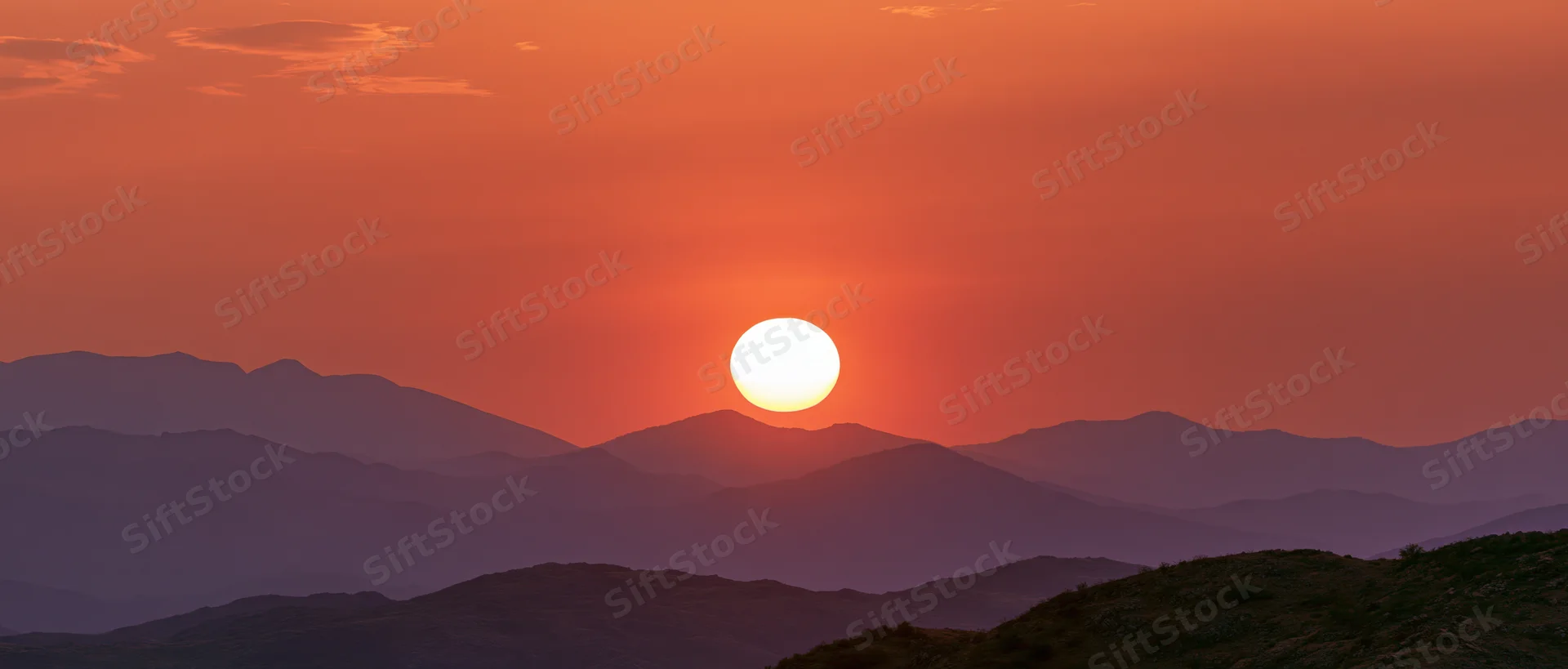 Large glowing sun at sunset above layered mountain silhouettes in a wide landscape view