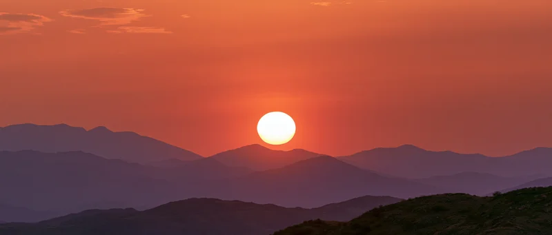 Large glowing sun at sunset above layered mountain silhouettes in a wide landscape view