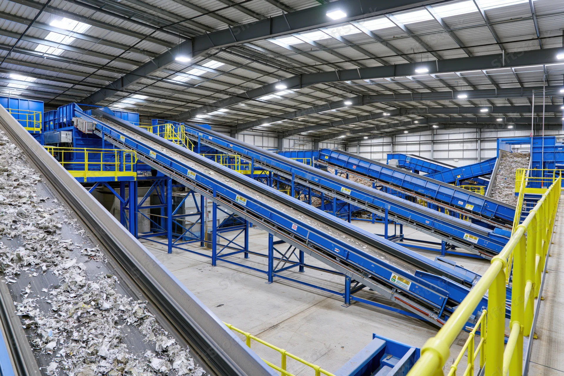 Industrial waste sorting facility interior with multiple conveyor belts transporting mixed debris across blue metal frameworks and yellow safety railings