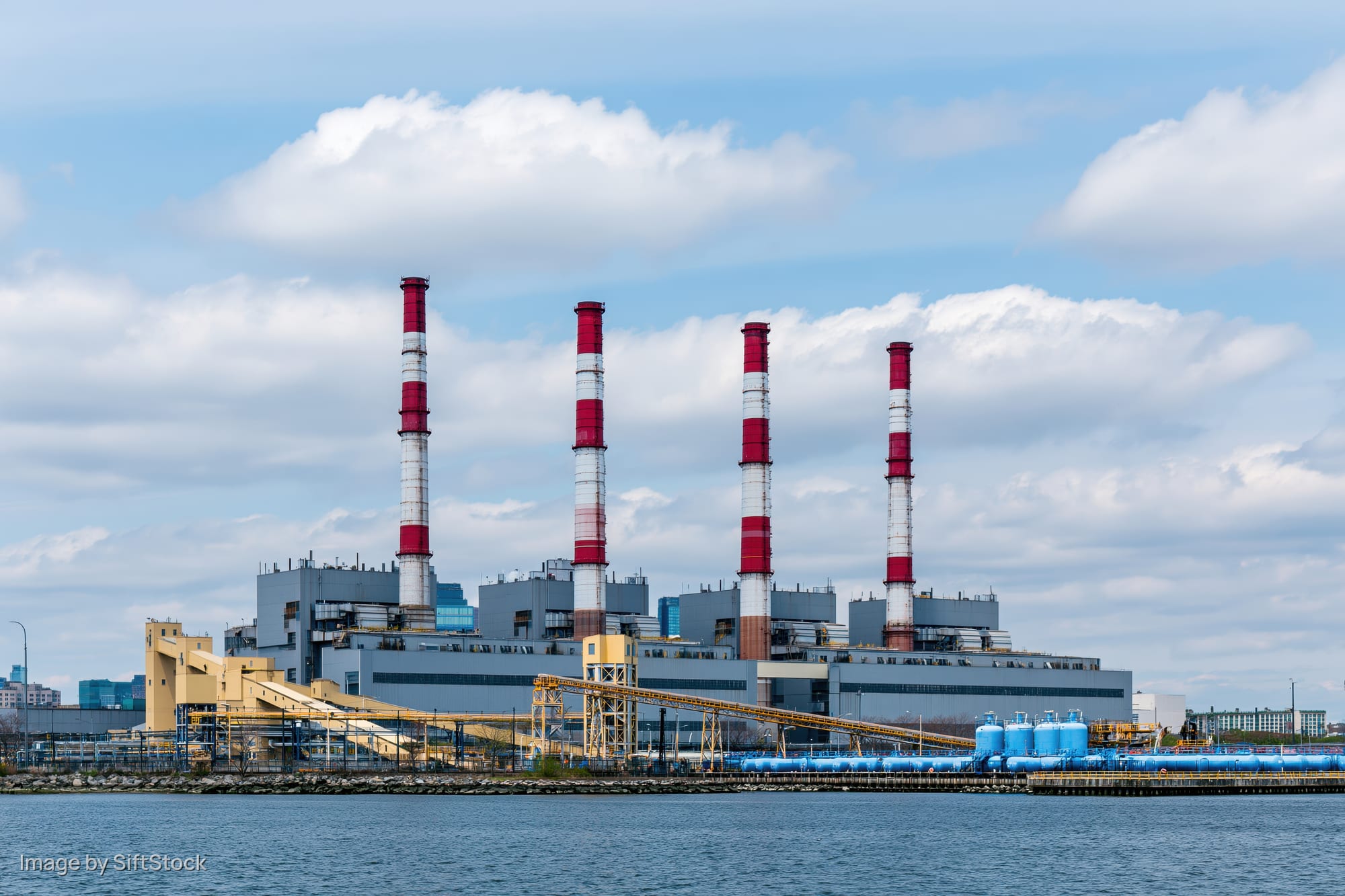 Large urban power plant with red-and-white smokestacks viewed across a river under a partly cloudy sky