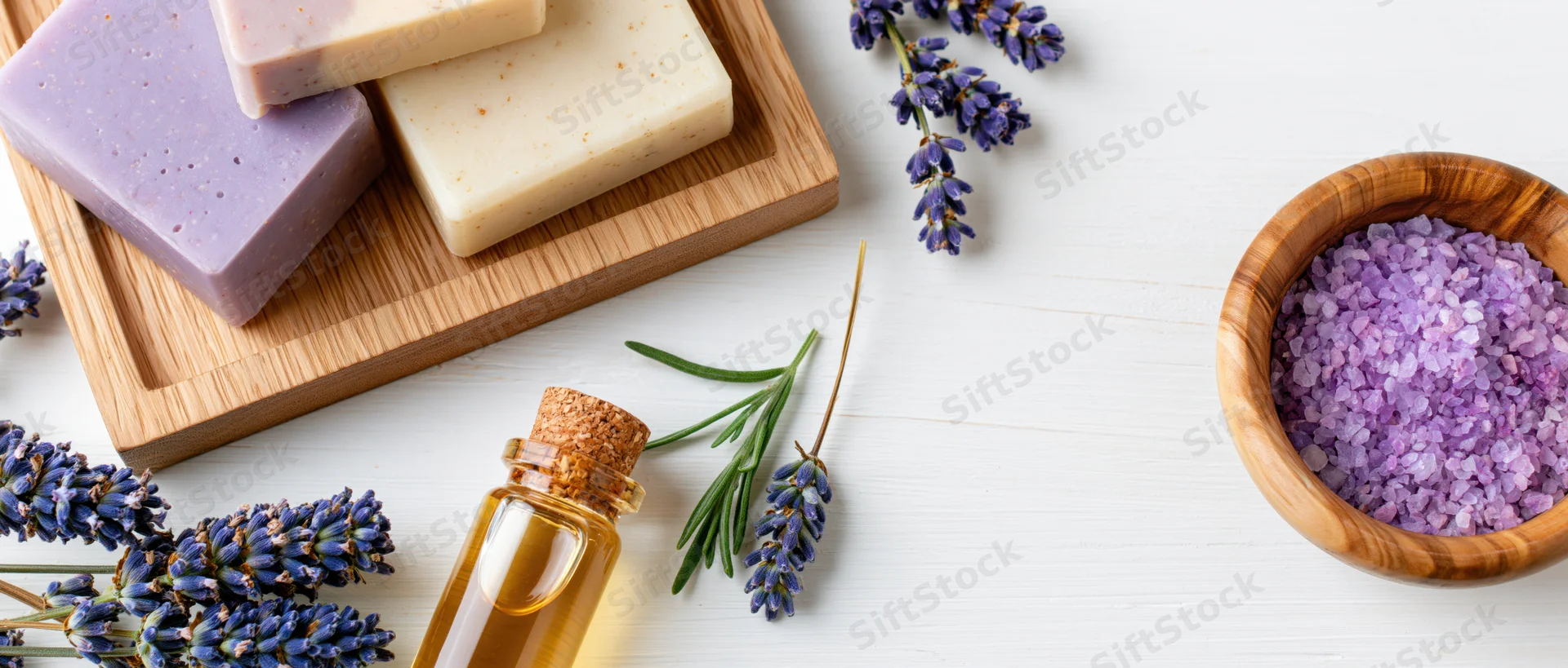 Lavender soap bars and essential oil arranged on wooden tray with flowers and bath salt on white surface