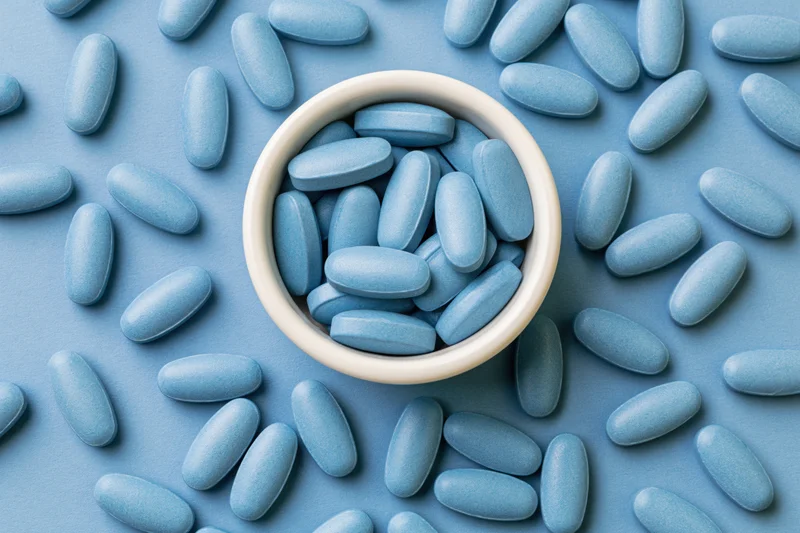 Blue oval supplement tablets scattered around a white bowl on a blue surface