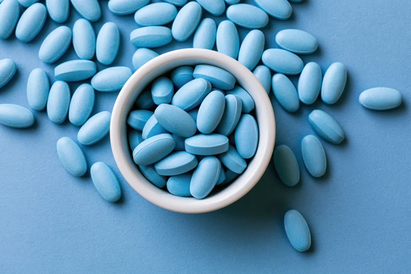 Light blue oval supplement tablets scattered across a flat blue surface with some in a white ceramic bowl