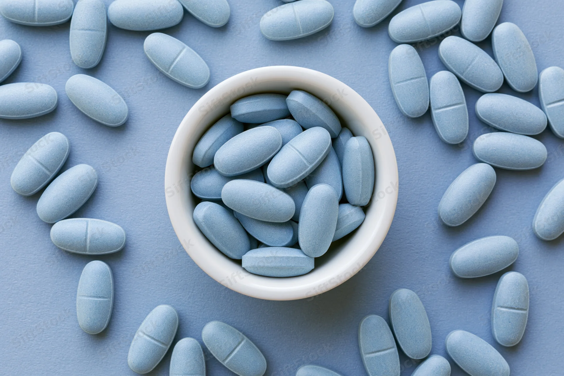 Light blue oval supplement tablets scattered around a white bowl on a flat blue surface