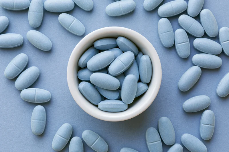Light blue oval supplement tablets scattered around a white bowl on a flat blue surface