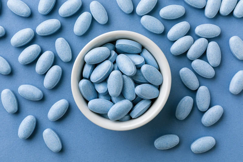 Blue oval supplement tablets scattered around a white bowl on a blue surface