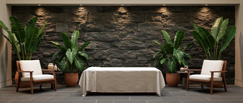 White-draped massage table between two wooden chairs with green plants against a stone wall in a spa treatment room