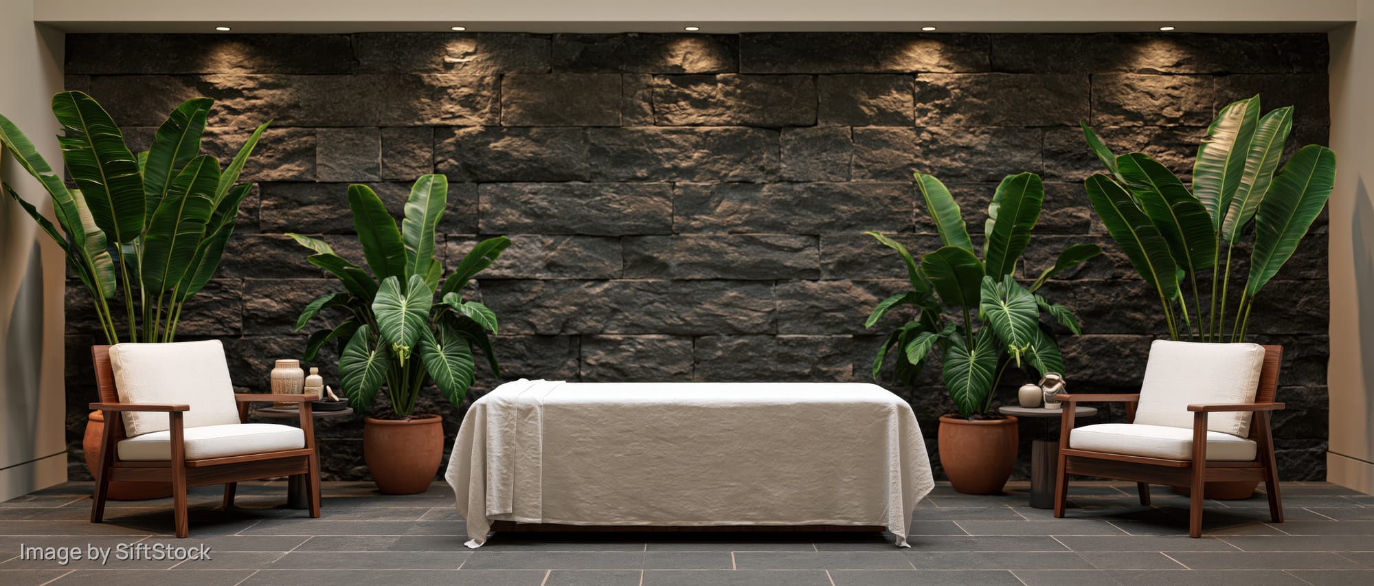 White-draped massage table between two wooden chairs with green plants against a stone wall in a spa treatment room