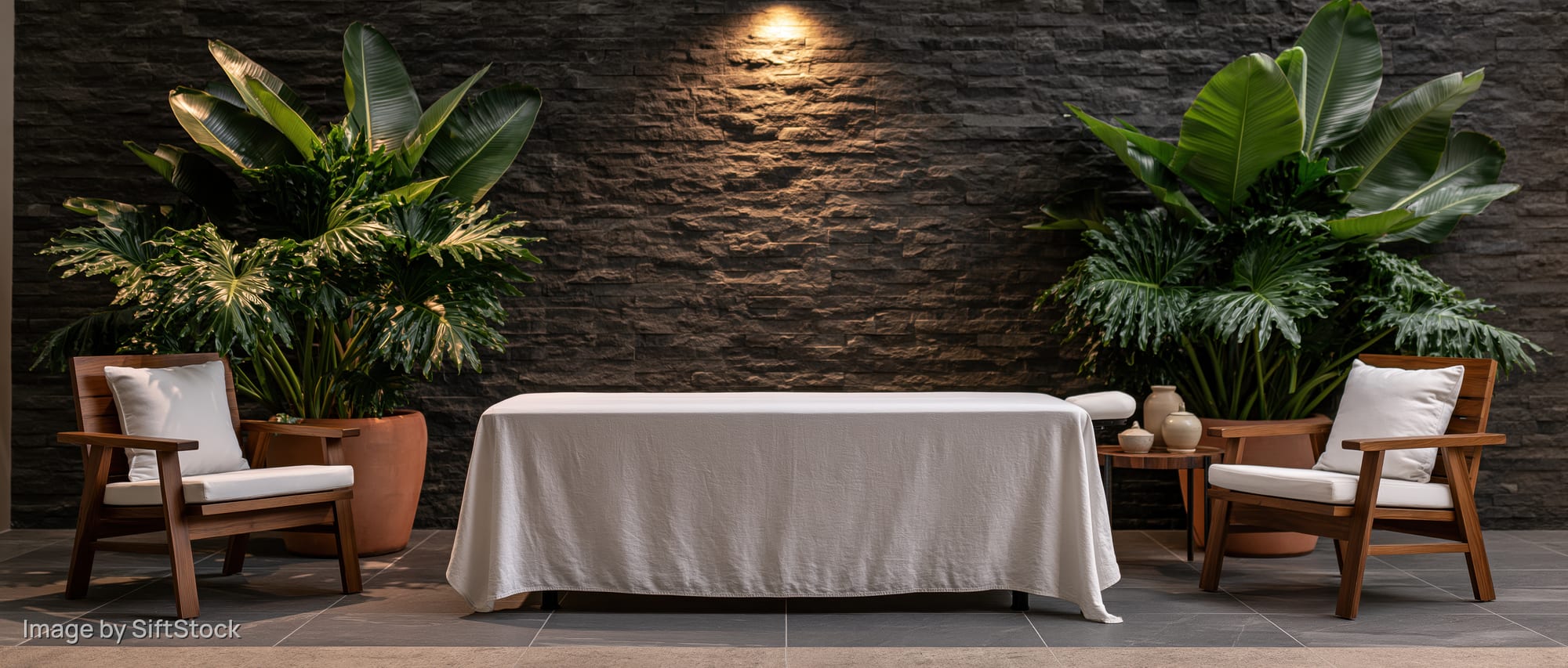 White-draped massage table between wooden chairs with tropical plants against stone wall in spa treatment room