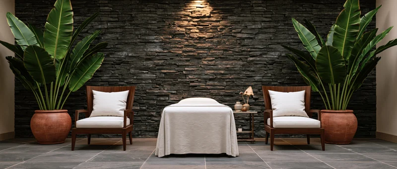 Massage table draped in white between two wooden chairs with potted banana plants against a stone wall in a spa treatment room