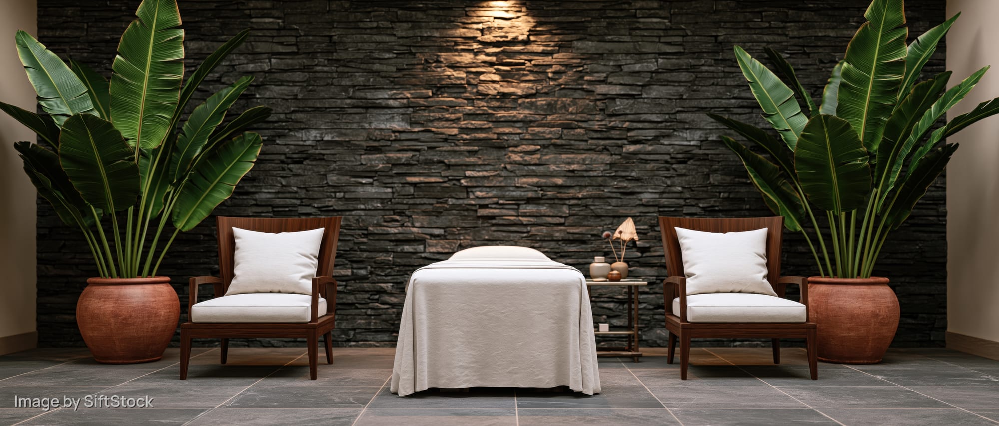Massage table draped in white between two wooden chairs with potted banana plants against a stone wall in a spa treatment room