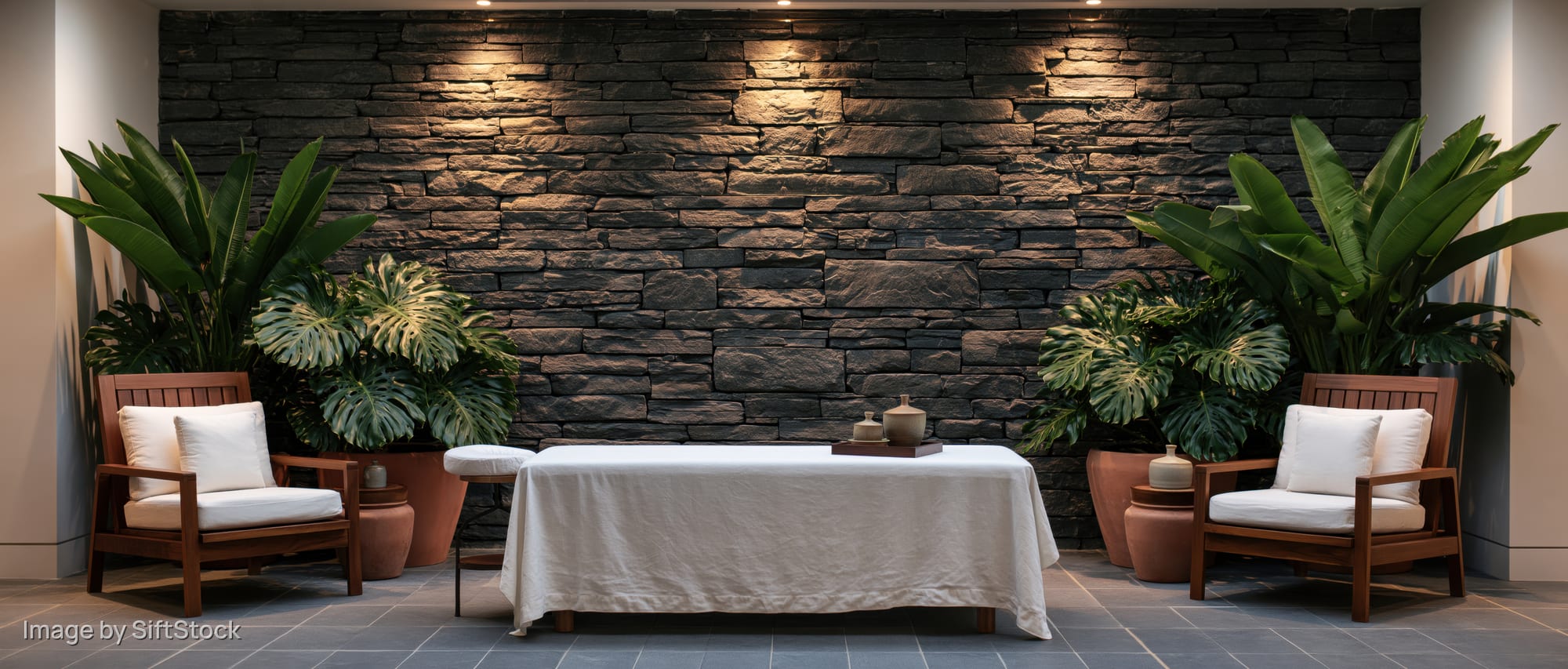 Massage table draped in white cloth between two wooden chairs with plants against a stone wall in a spa treatment room
